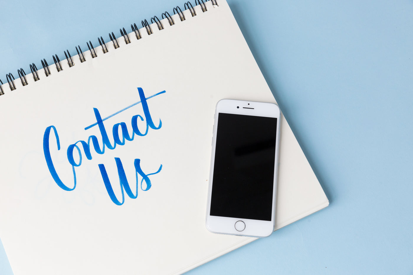 An iPhone is sitting on top of a spiral-bound notepad with "Contact Us" printed on it.