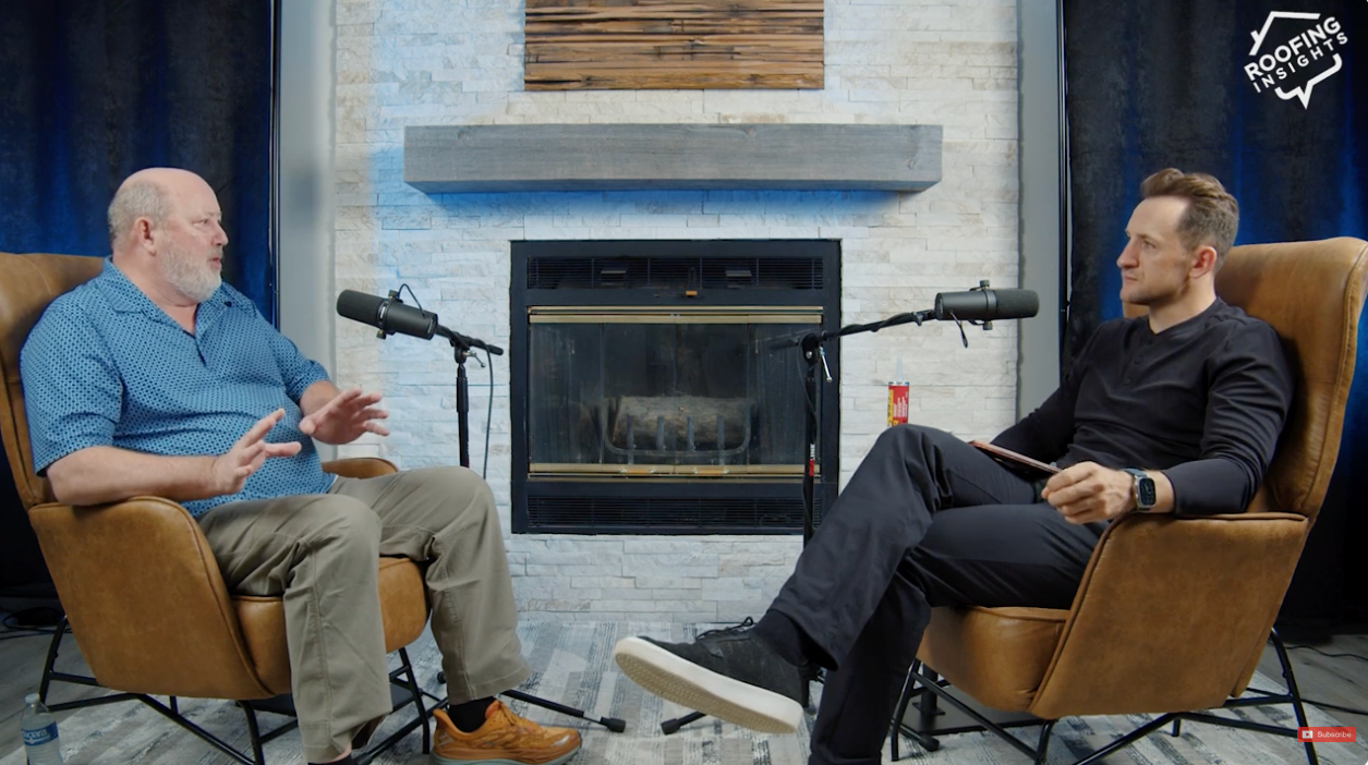 Two men sitting in chairs in a studio setting with microphones and a fireplace.