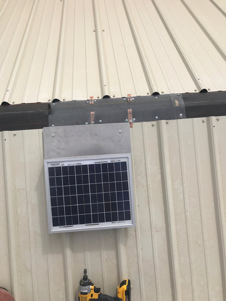 A Solar RidgeBlaster is being installed on a beige shed's metal roof. Shows the open ridge with netting covering over the fan unit to keep pests out.