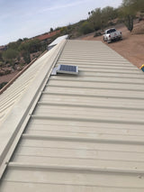 Solar RidgeBlaster has been installed on a beige metal roof of a shed in the desert with a truck and a house in the background.