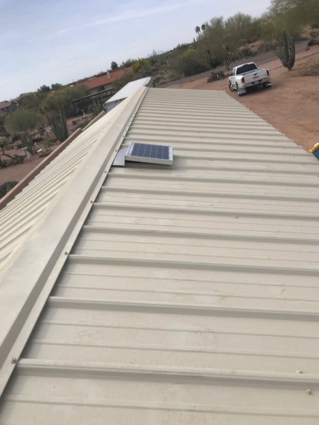 Solar RidgeBlaster has been installed on a beige metal roof of a shed in the desert with a truck and a house in the background.
